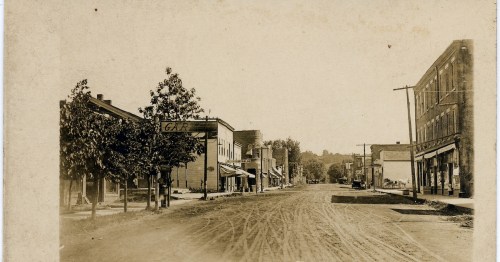 A vintage street scene featuring buildings, trees, and dirt roads, evoking a historic small-town atmosphere.