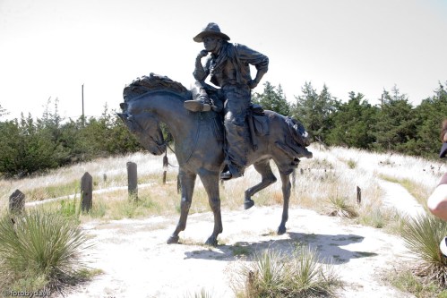 A bronze statue of a cowboy on horseback, thoughtfully resting his chin on one hand, surrounded by grass and trees.