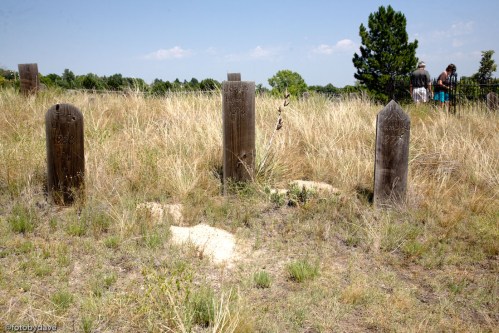 Four wooden grave markers stand in a grassy field, with people in the background near a fence.