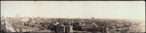 Panoramic view of a city skyline, showcasing buildings, streets, and a hazy atmosphere in the distance.