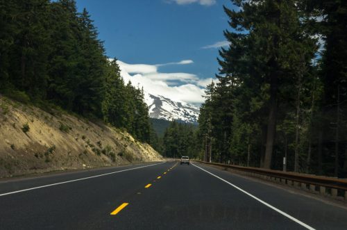 A scenic highway lined with tall trees, leading to snow-capped mountains under a blue sky with clouds.