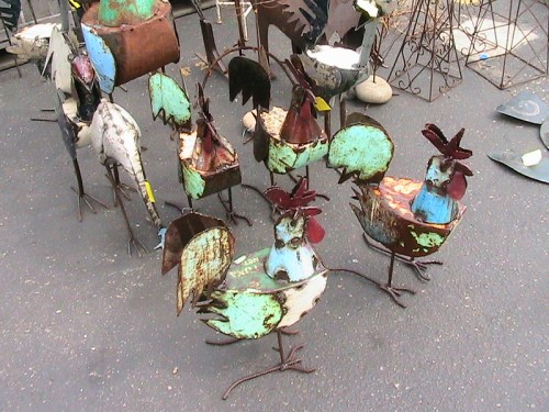 A group of colorful metal chickens with rusted finishes, displayed outdoors on a paved surface.