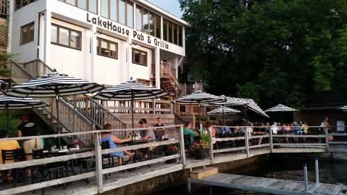 A lakeside pub with outdoor seating under striped umbrellas, surrounded by greenery and a wooden dock.