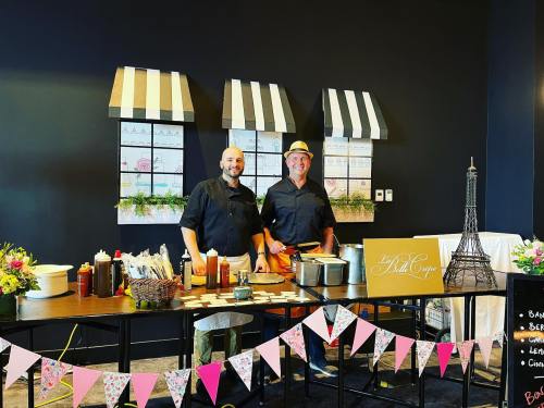 Two chefs stand behind a table with crepe-making supplies, decorated with flowers and a Parisian theme backdrop.