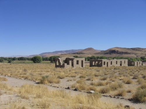 Desert landscape with ruins of adobe buildings, surrounded by dry grass and distant mountains under a clear blue sky.