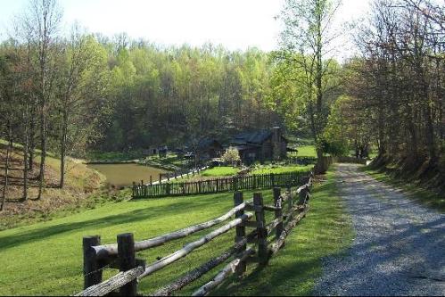 A gravel path leads to a rustic house by a pond, surrounded by lush green trees and a wooden fence.