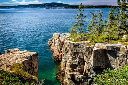 A scenic view of rocky cliffs and clear blue water, surrounded by lush green trees and distant hills.