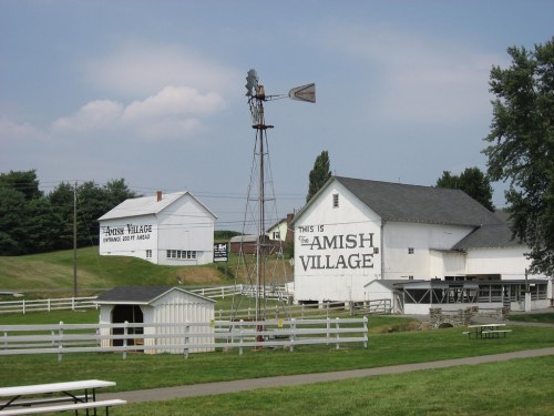 A rural scene featuring white buildings with "This is the Amish Village" sign, a windmill, and green grass.