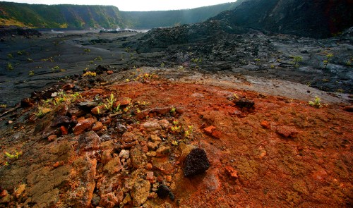 A volcanic landscape with black lava rock and patches of red soil, surrounded by steep, green hills in the background.