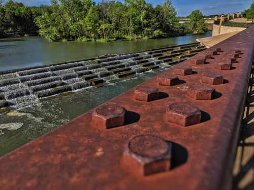 A close-up of a rusted metal railing with large bolts, overlooking a river with a small waterfall and greenery in the background.