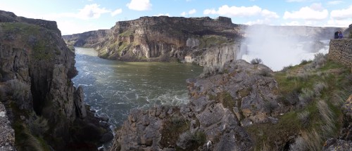A panoramic view of a river canyon with rocky cliffs and a waterfall, under a blue sky with scattered clouds.