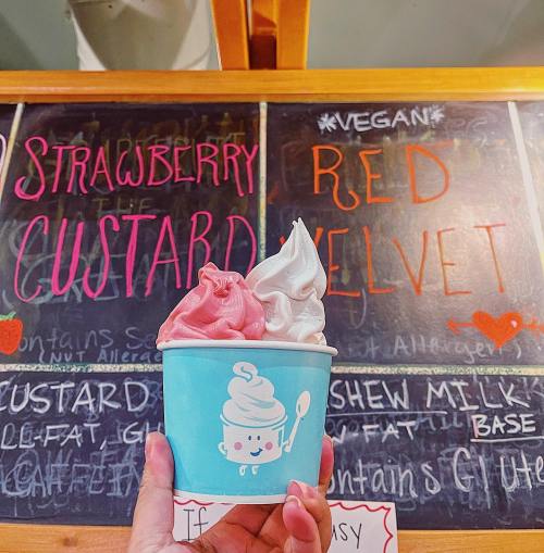 A hand holding a cup of strawberry custard and red velvet soft serve in front of a colorful menu board.