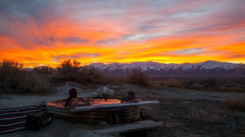 Two people relax in a hot tub at sunset, with vibrant orange and purple skies and snow-capped mountains in the background.