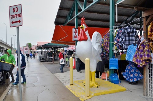 A large chicken statue stands on a sidewalk near a market, with people and stalls visible in the background.