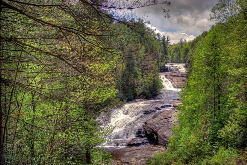 A scenic view of a waterfall surrounded by lush green trees and rocky terrain under a partly cloudy sky.