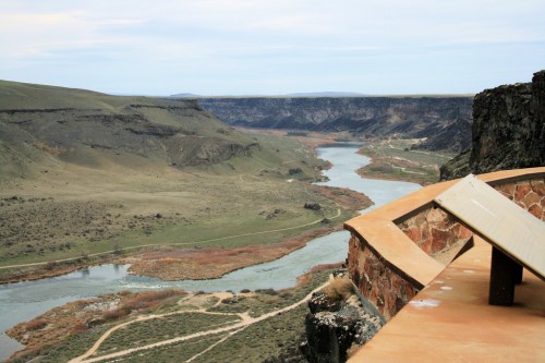 A scenic view of a winding river surrounded by green hills and rocky cliffs under a cloudy sky.