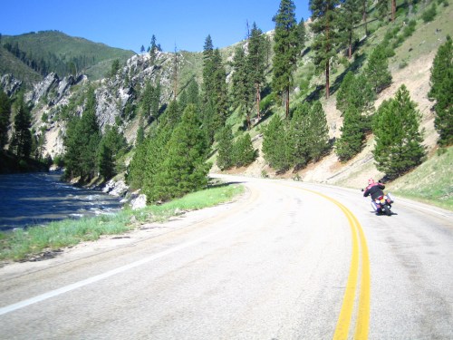 A motorcyclist rides along a winding road beside a river, surrounded by lush green trees and mountains.
