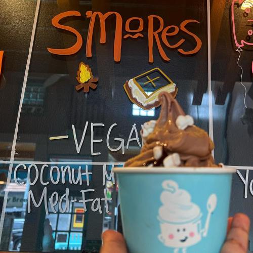 A hand holds a cup of chocolate soft serve topped with mini marshmallows, with a colorful menu in the background.