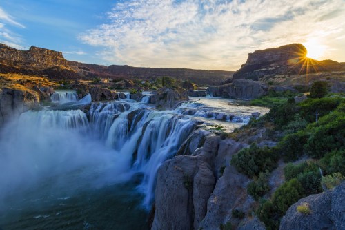 A stunning waterfall cascades over rocky cliffs under a vibrant sky at sunset, surrounded by rugged terrain.