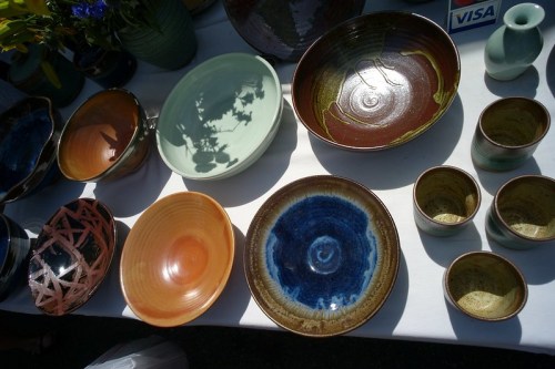 A variety of colorful pottery, including bowls and cups, displayed on a table with flowers in the background.