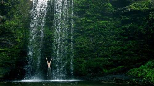 A person stands under a waterfall, arms raised, surrounded by lush green cliffs and flowing water.