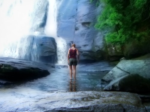 A person stands in shallow water near a waterfall, surrounded by rocks and lush greenery.