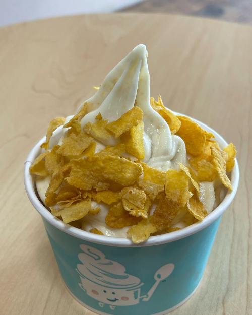 A cup of soft-serve ice cream topped with crunchy cornflakes, set on a wooden surface.