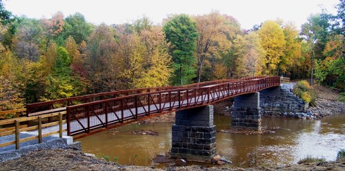 A metal bridge spans a calm river, surrounded by vibrant autumn trees in shades of yellow, orange, and green.