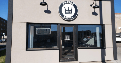 Exterior of Empire Pizza with a round logo, "Open" sign, and large windows on a sunny day.