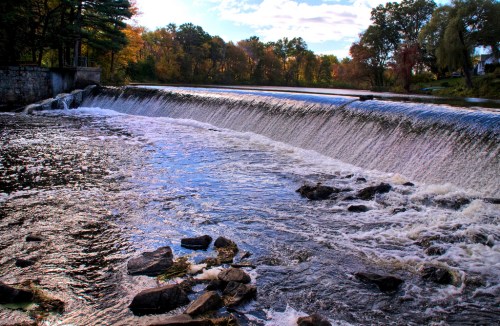 A serene waterfall cascades over rocks, surrounded by autumn foliage and a clear blue sky.
