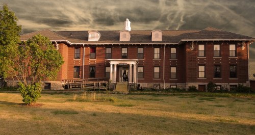 A large, historic brick building with a porch, surrounded by grass under a dramatic, cloudy sky.