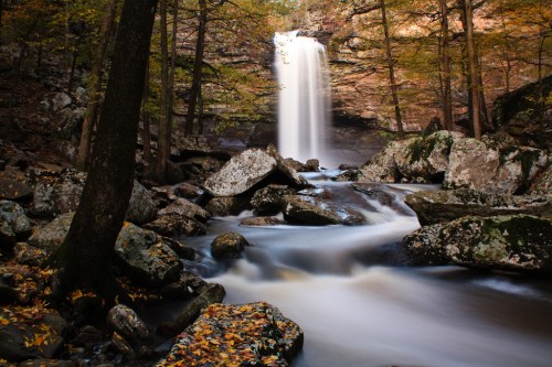 A serene waterfall cascades over rocks, surrounded by autumn foliage and a tranquil stream.