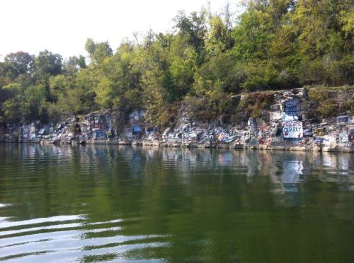 A rocky shoreline covered in graffiti, reflecting in calm water surrounded by trees.