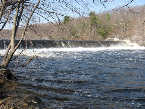A serene river scene featuring a waterfall cascading over a dam, surrounded by trees and a clear blue sky.