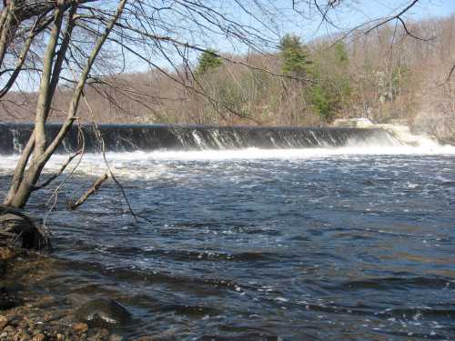 A serene river scene featuring a waterfall cascading over a dam, surrounded by trees and a clear blue sky.