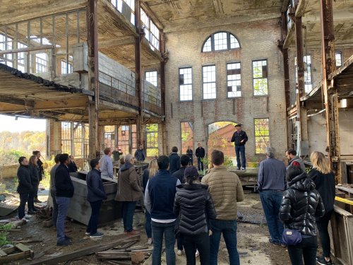 A group of people gathers in an abandoned industrial building, listening to a speaker in a sunlit, open space.