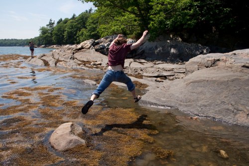 A person jumps over a rocky area by the water, while another person stands in the background near the shore.