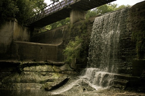 A serene waterfall cascades over a stone wall beneath a wooden bridge, surrounded by lush greenery.