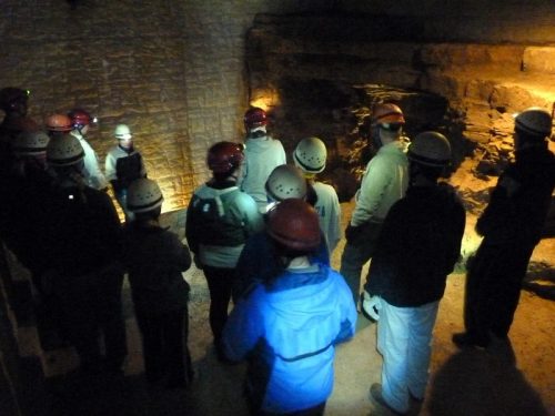 A group of people in helmets listens to a guide in a dimly lit cave setting.