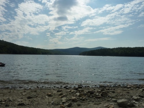 A serene lake surrounded by hills, with a rocky shore and a cloudy sky reflecting on the water's surface.