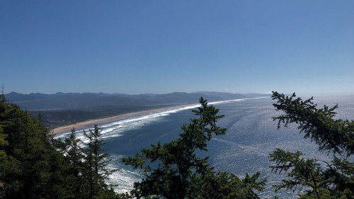 A panoramic view of a coastline with waves, sandy beach, and mountains under a clear blue sky, framed by trees.