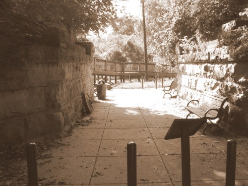 A sepia-toned pathway leads to a park area with benches and trees, framed by stone walls.