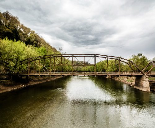 A weathered metal bridge spans a calm river, surrounded by lush greenery under a cloudy sky.