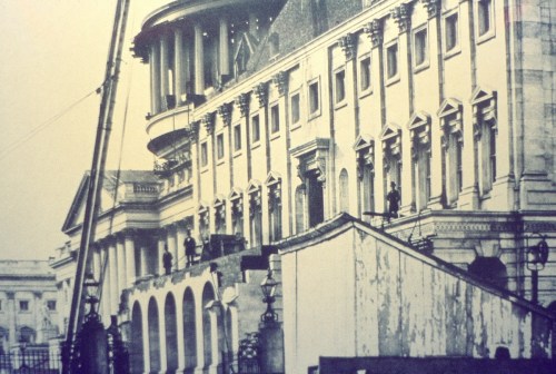 Historic building with a crane, featuring ornate architecture and workers on the upper levels. Black and white photo.