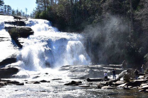 A scenic waterfall cascades over rocks, with mist rising and people enjoying the view nearby.