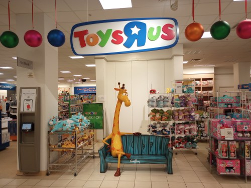Toys "R" Us store entrance featuring a giraffe statue and colorful decorations, with shelves of toys in the background.