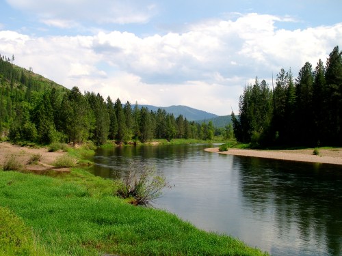 A serene river flows through a lush green landscape, surrounded by trees and mountains under a partly cloudy sky.