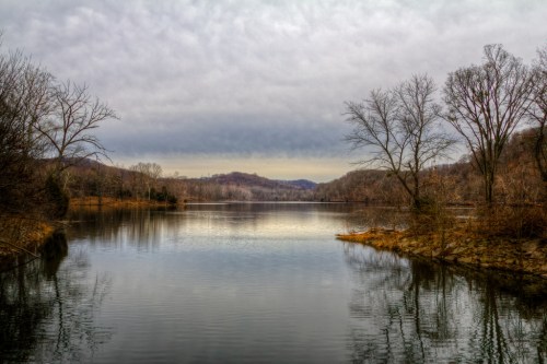 A tranquil lake surrounded by bare trees and rolling hills under a cloudy sky, reflecting the serene landscape.