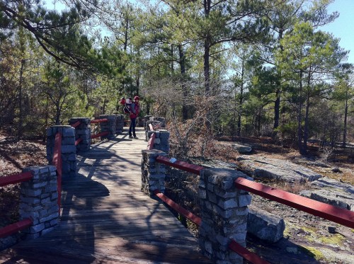A wooden bridge with stone railings in a forested area, with two people standing on it surrounded by trees.