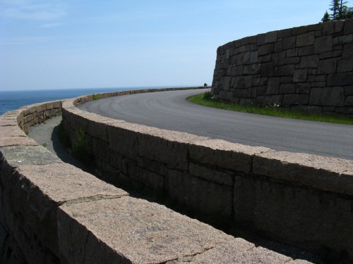 A curved stone wall along a coastal road, with the ocean visible in the background under a clear blue sky.